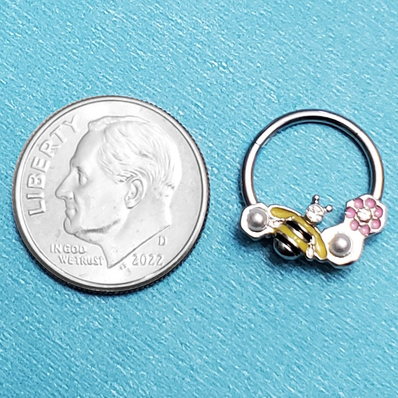 A surgical steel bee septum ring pictured on an aqua background next to a dime for a size comparison.