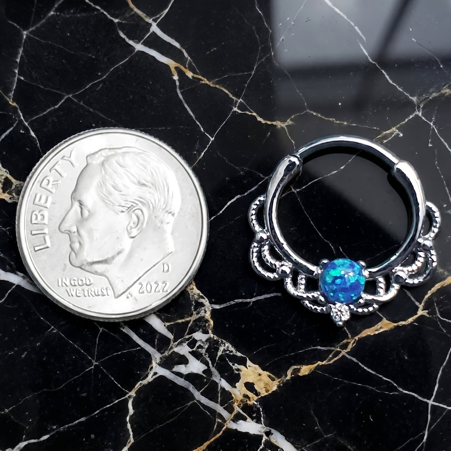 Filigree Blue Opal Septum Clicker 16 Gauge 316L Surgical Steel Brass pictured next to a dime on a black marble countertop to show a size comparison.