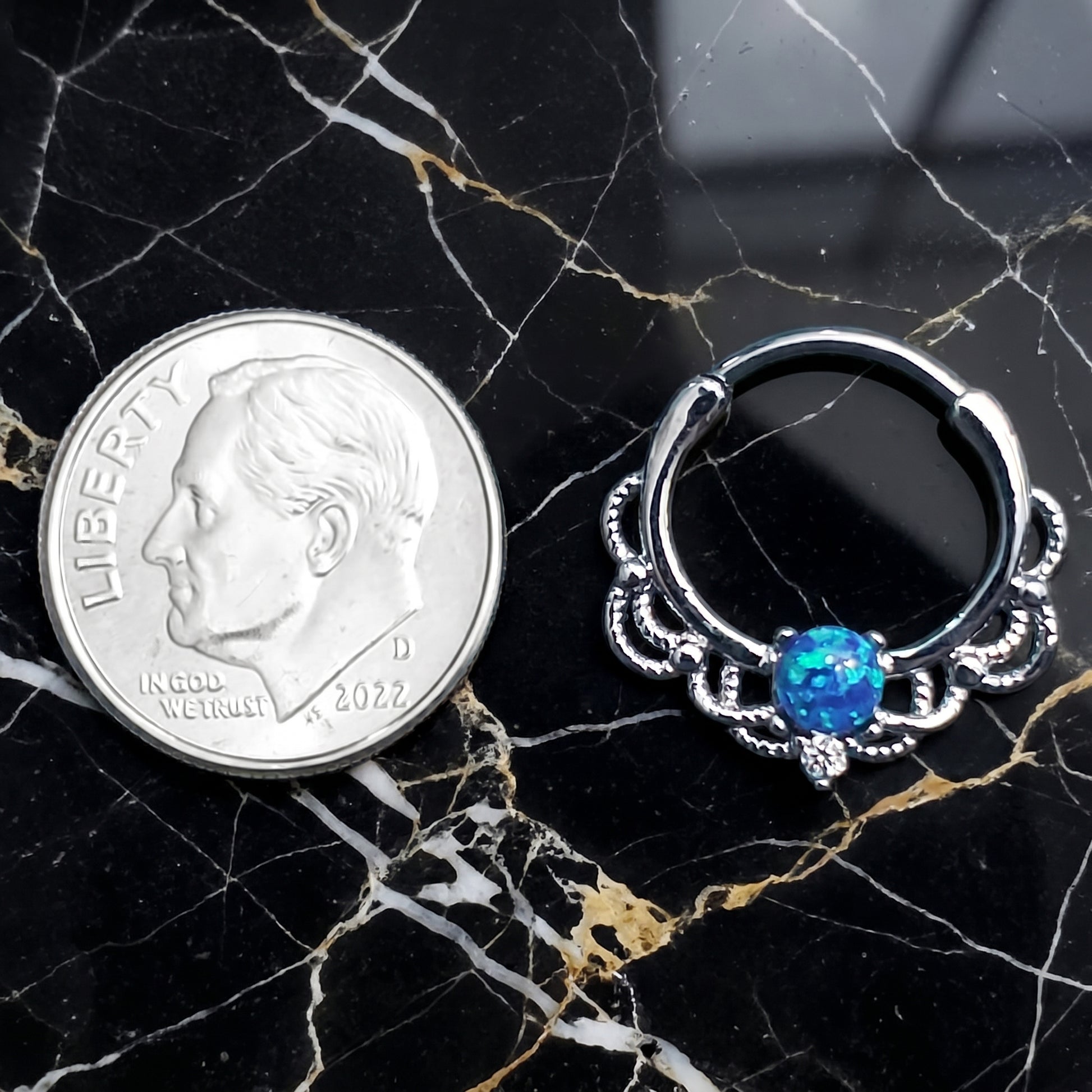 Filigree Blue Opal Septum Clicker 16 Gauge 316L Surgical Steel Brass pictured next to a dime on a black marble countertop to show a size comparison.