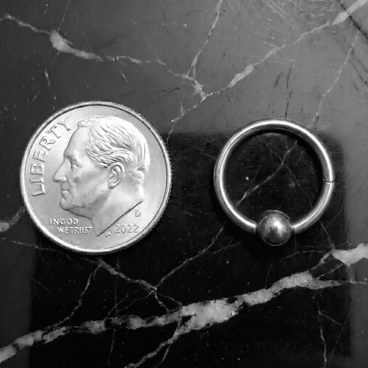 Hinged Segment Fixed Captive Bead Ring 16 Gauge 316L Surgical Steel pictured next to a US dime on a black marble countertop to show a size comparison.