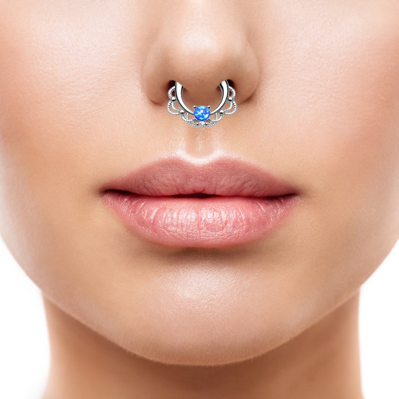 A surgical steel opal septum clicker featuring a blue opal centered against a lacy design with the piece pictured in the septum piercing of a female.