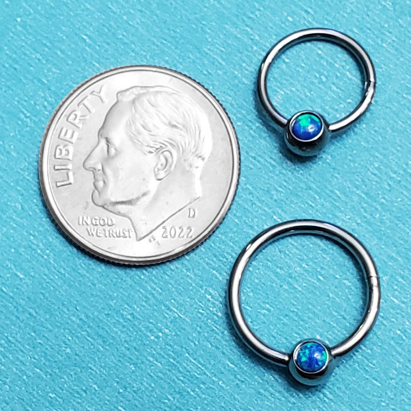 Two sizes of surgical steel opal septum rings pictured on an aqua background next to a dime for a size comparison.