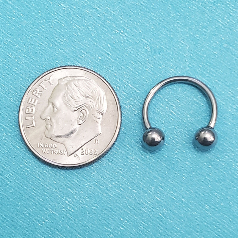 A surgical steel septum ring pictured on an aqua background next to a dime for a size comparison.