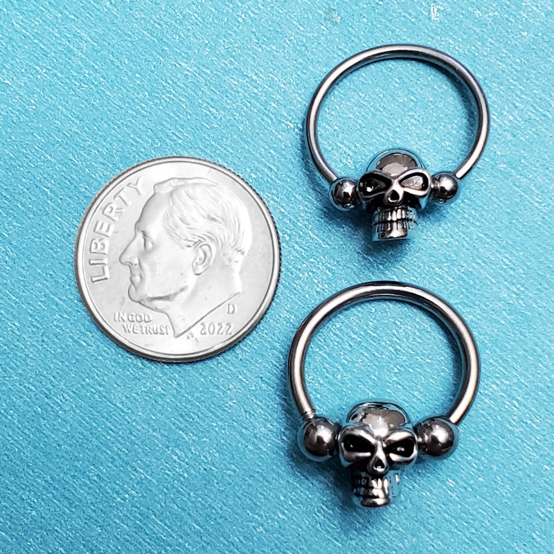 Two sizes of surgical steel skull septum rings pictured on an aqua background next to a dime for a size comparison.