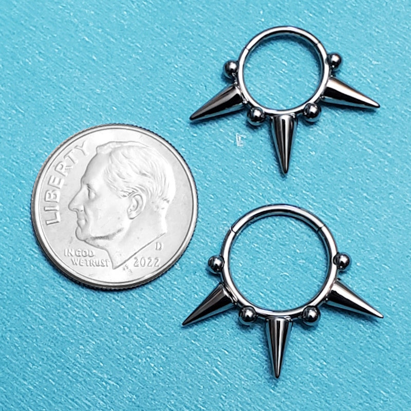 A surgical steel spiked septum ring pictured on an aqua background next to a dime for a size comparison.