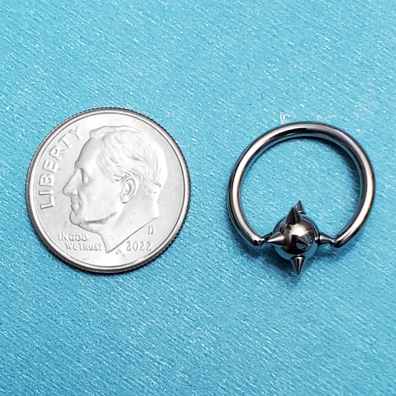 A surgical steel spiky septum ring pictured on an aqua background next to a dime for a size comparison.