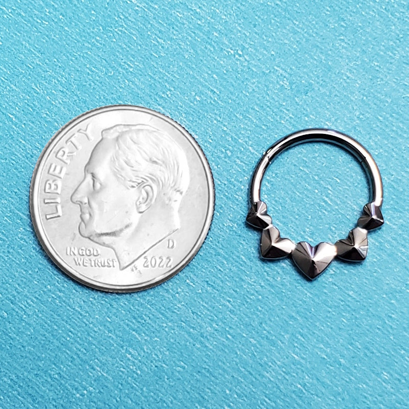 A titanium heart septum ring pictured on an aqua background next to a dime for a size comparison.