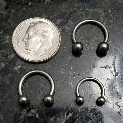Three sizes 18G, 16G, 14G titanium horseshoe septum rings pictured next to a dime for a size comparison pictured on a black marble countertop.