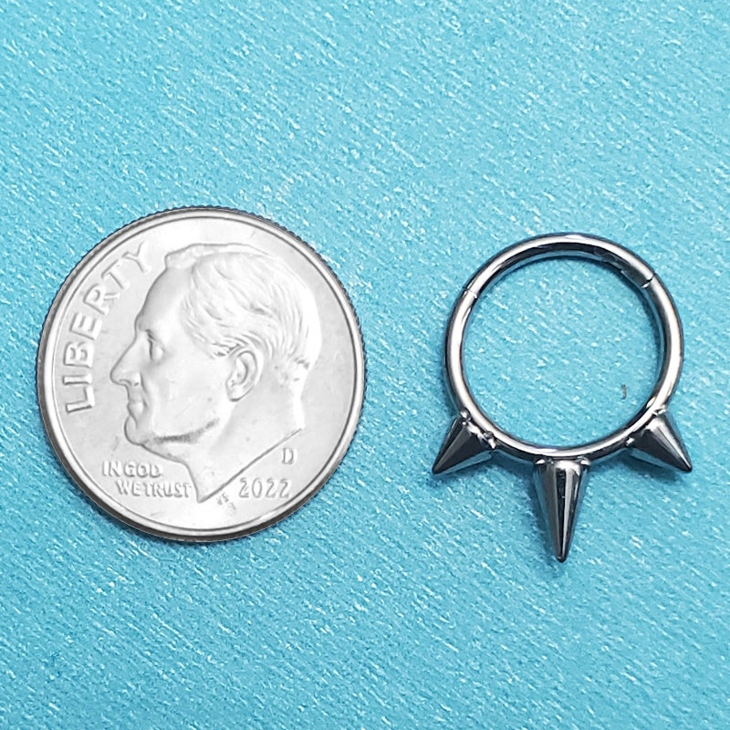 A titanium septum ring with spikes pictured on an aqua background next to a dime for a size comparison.