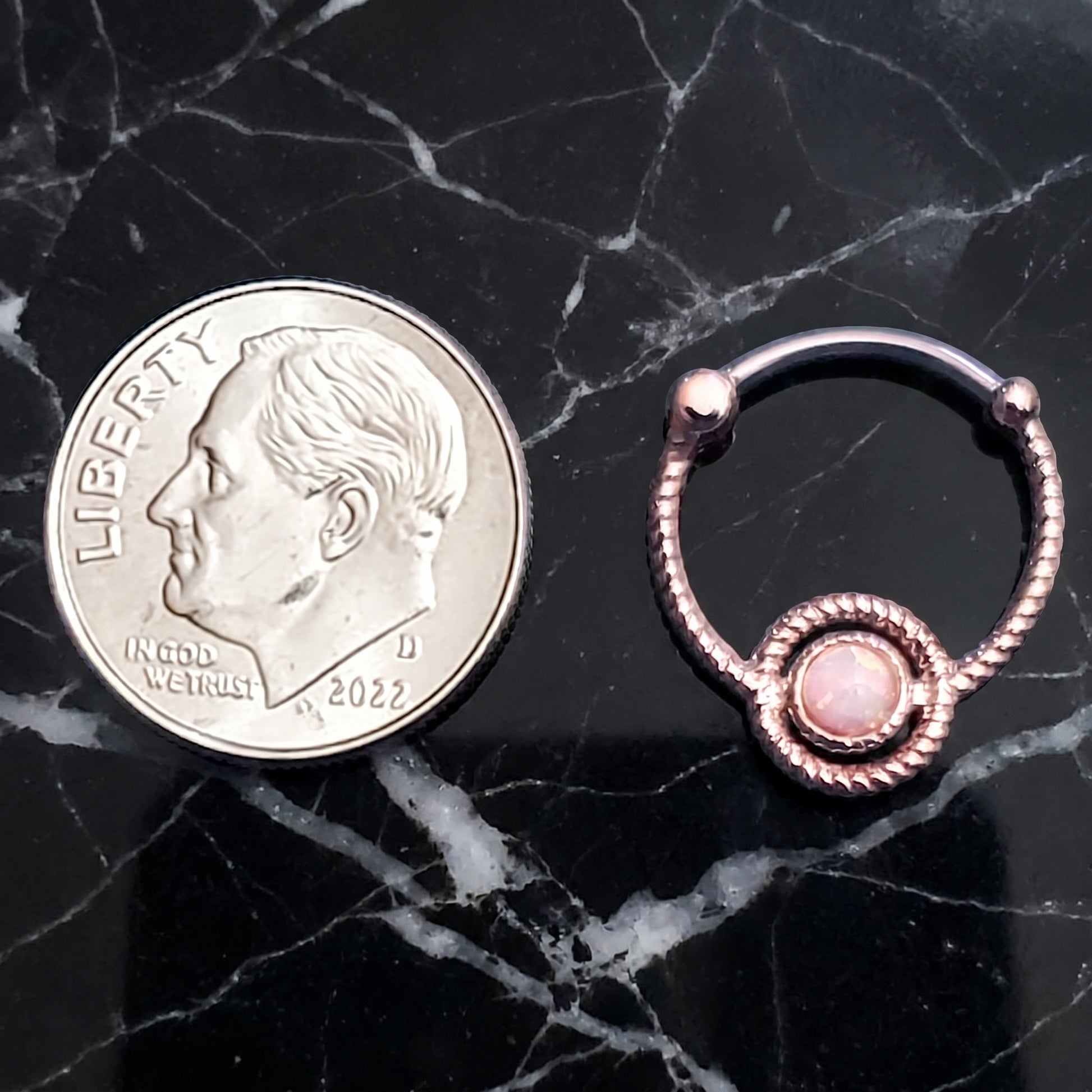 A twisted rope rose gold opal septum clicker pictured next to a dime on a black marble countertop to show the size comparison.